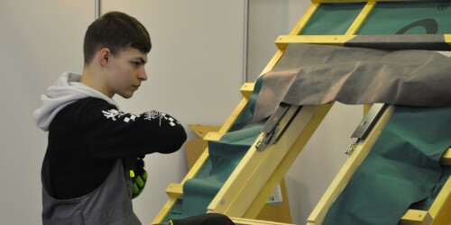 Uczeń ubrany w strój roboczy stojący przy makiecie do nauki układania dachówki